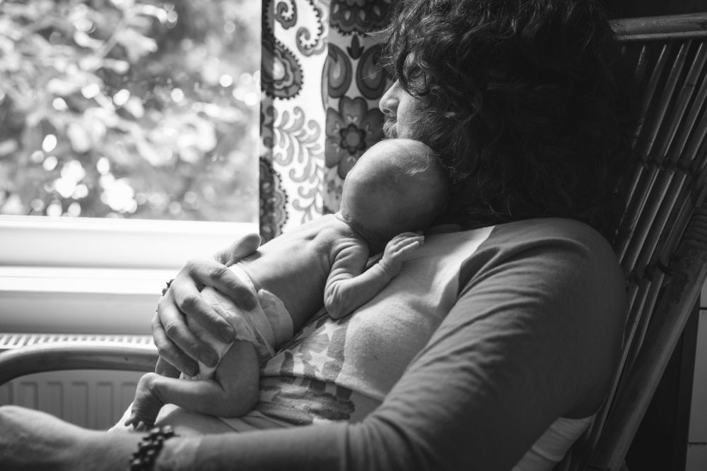 A tender black and white photo of a baby sleeping on an adult's chest in a rocking chair by a window.