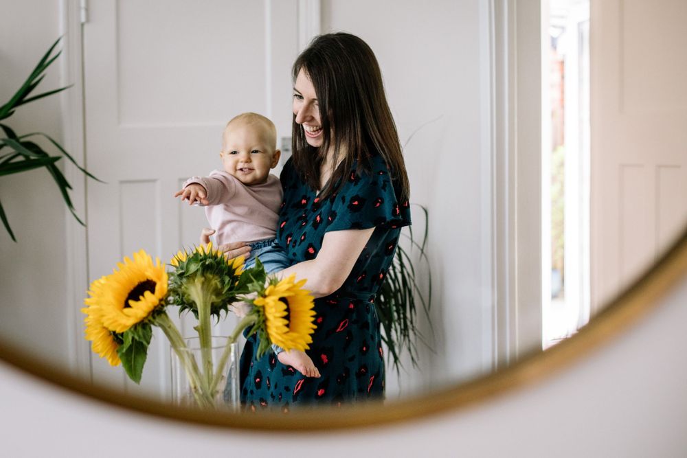 A reflection in a mirror shows a parent and baby next to bright yellow sunflowers in a bright modern interior.