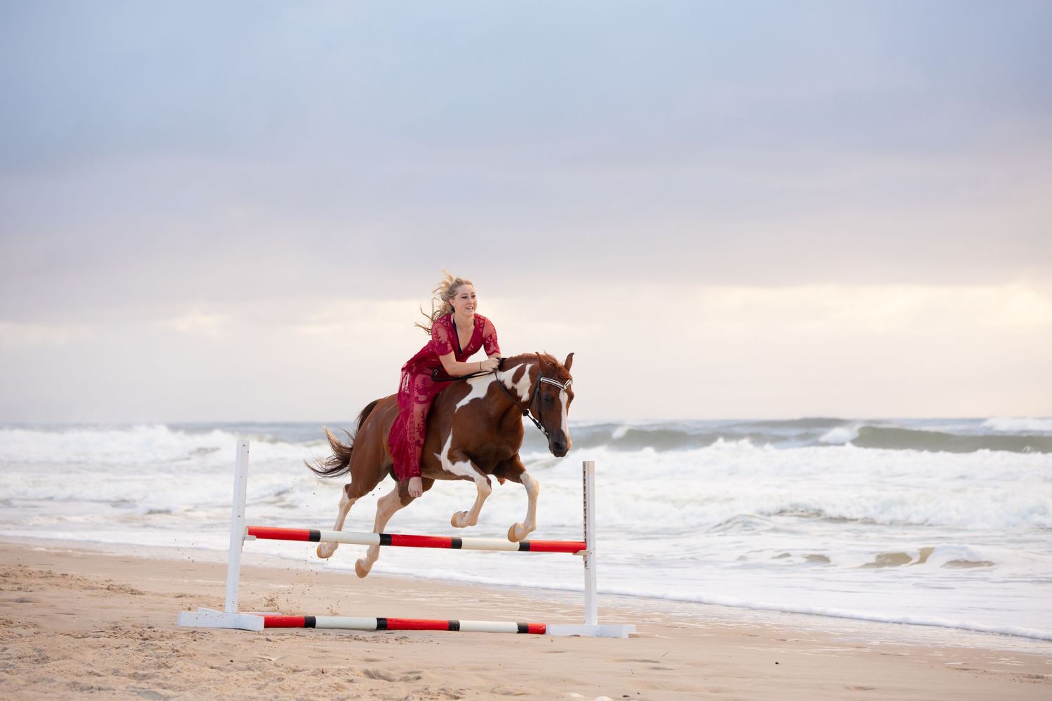 Young girl in red dress jumping a chestnut paint horse at the beach