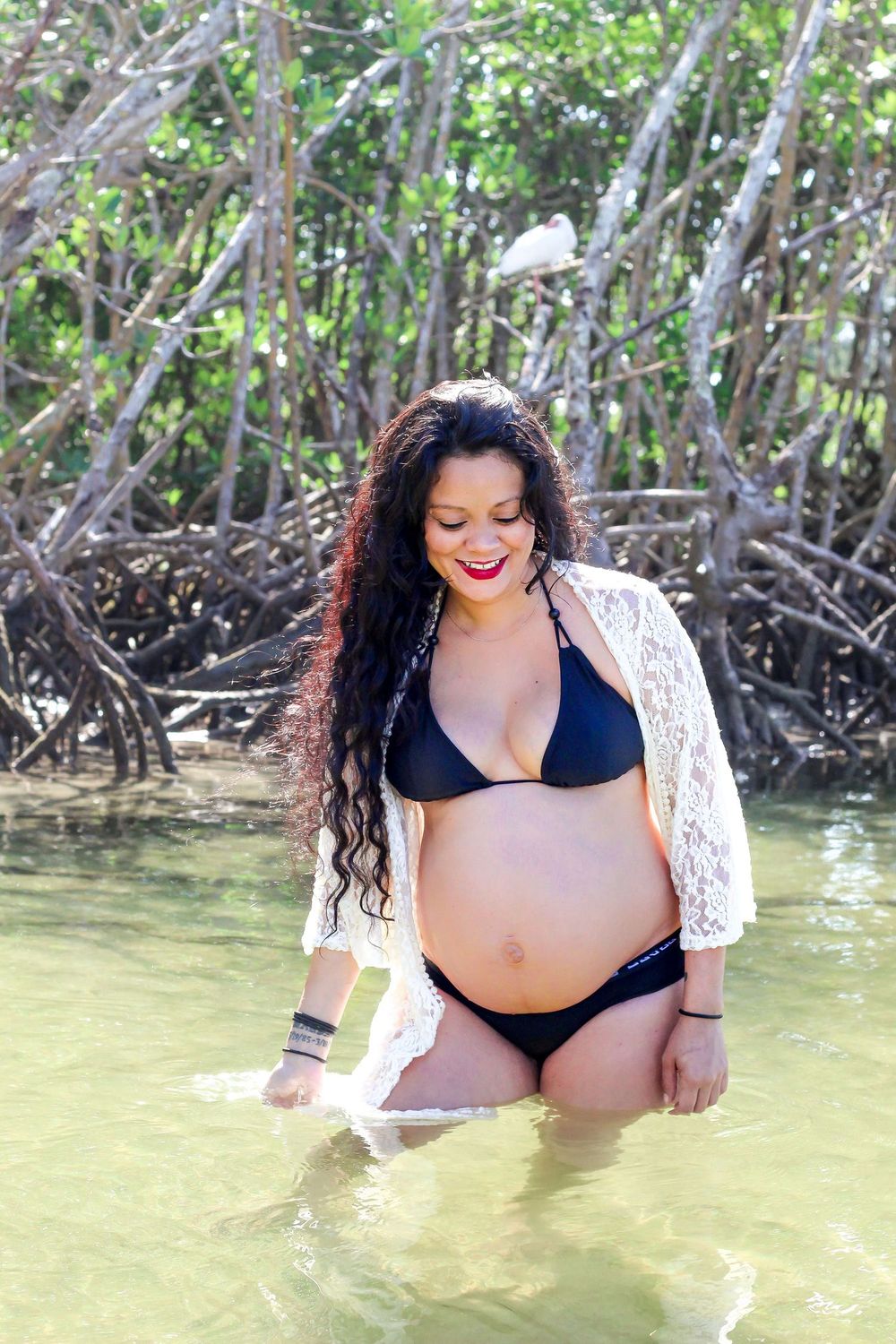 Pregnant woman in black bikini and white lace cover-up standing in shallow water near mangrove trees at dubios beach.