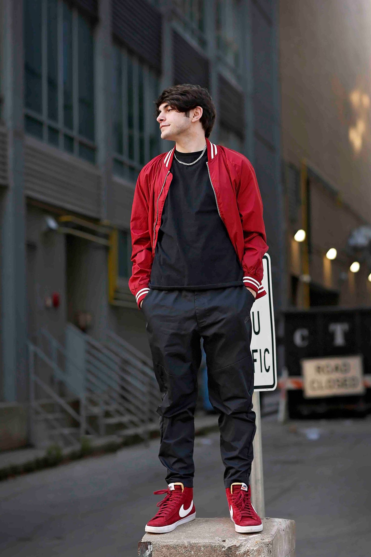Young man in red jacket and black pants poses on concrete barrier in urban setting.