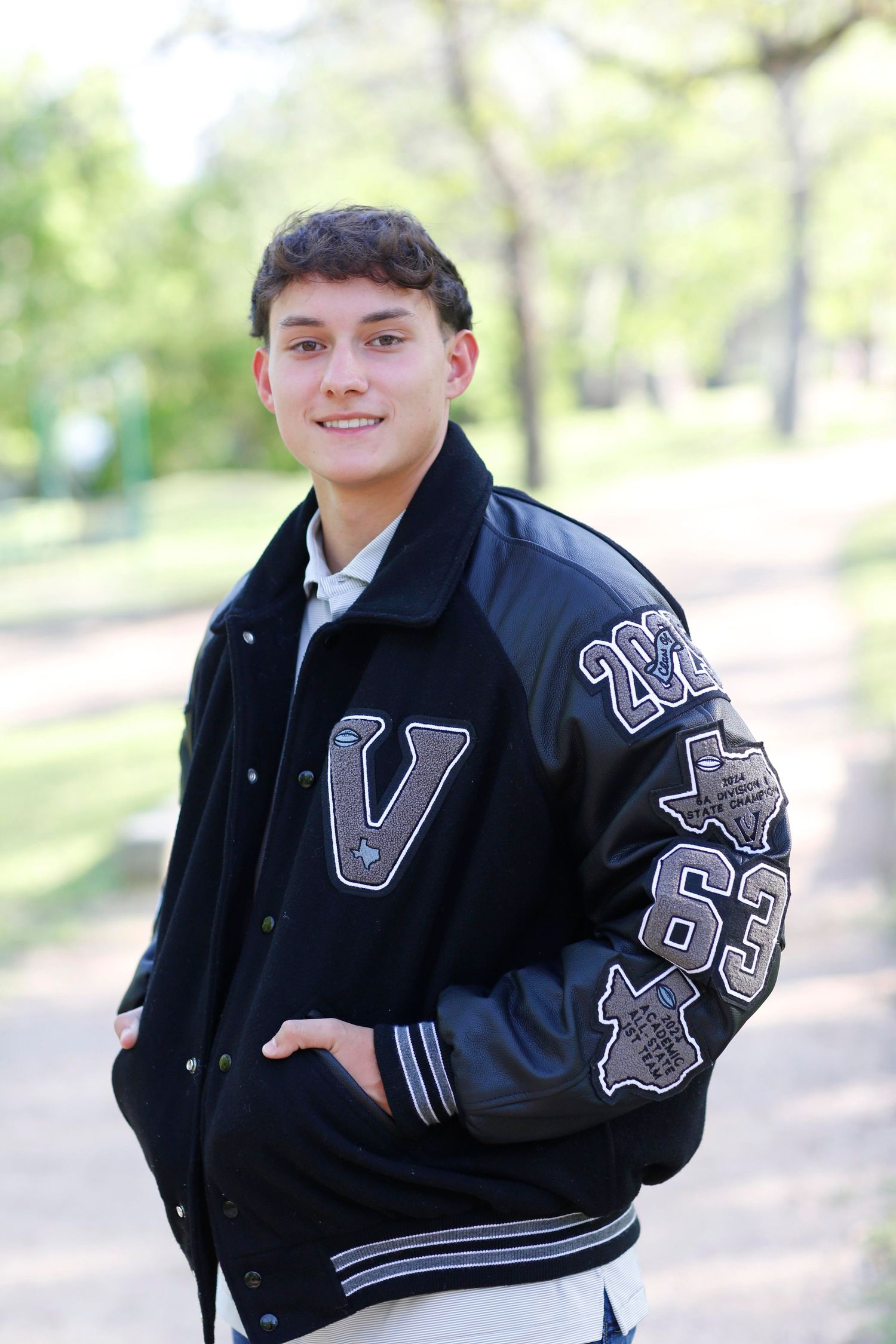 Person wearing varsity letterman jacket poses outdoors in park setting.