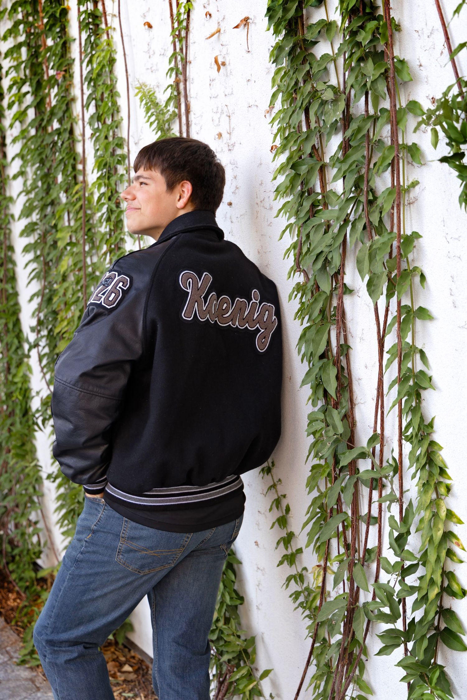 Person wearing a black varsity jacket and jeans poses against a wall with hanging green vines.