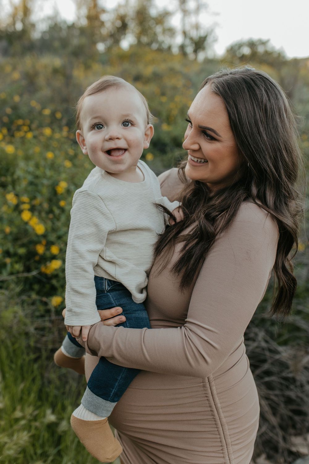 A mother holds her son while smiling in front of yellow wildflowers.