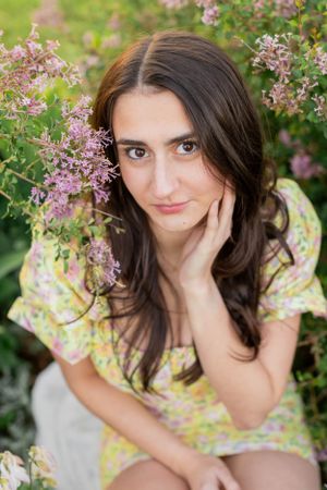 Senior girl session with girl sitting in a spring garden scene with blooming lilac bushes and soft natural lighting in Springfield, Missour.