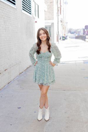 Senior session with girl in sage green mini dress and white boots poses on urban sidewalk with hands on hips in Springfield, Missouri.
