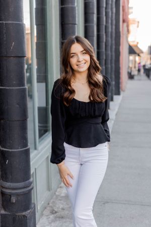 Senior session with girl in black blouse and white pants poses by building with black columns in Springfield, Missouri.