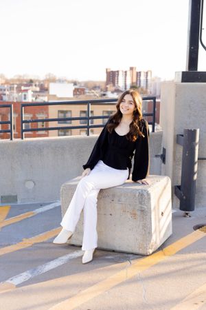 Senior session with girl in black top and white pants sits on concrete block on rooftop with city skyline view in Springfield, Missouri.