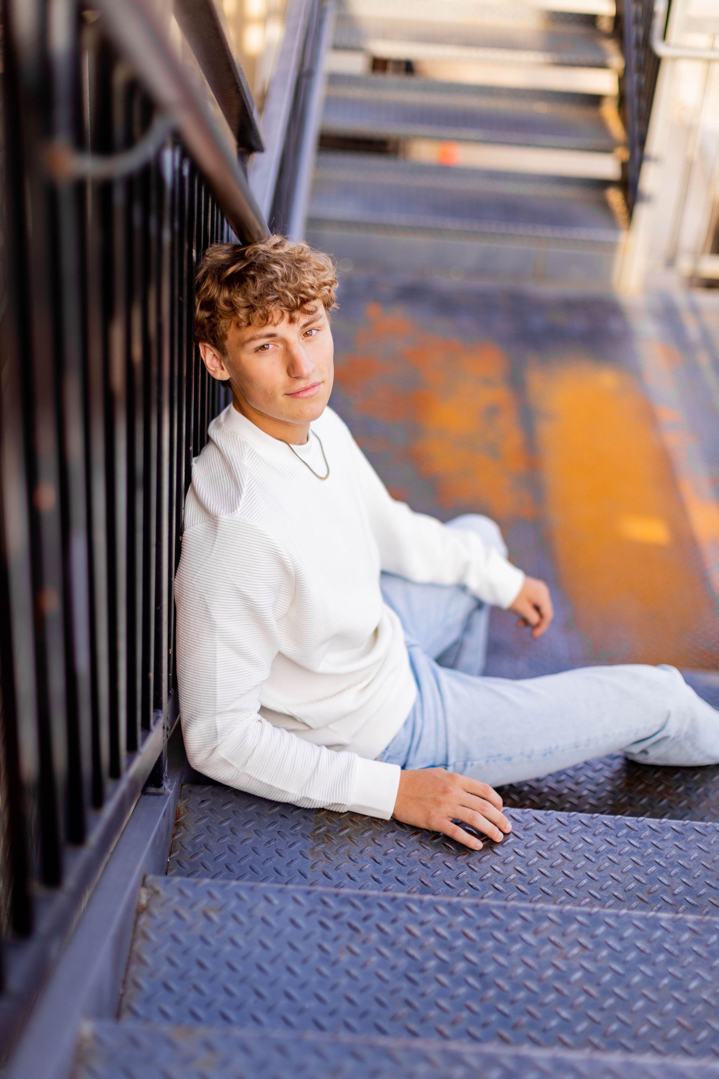 Senior session with boy in white sweater and jeans sits casually on metal industrial stairs in Springfield, Missouri.