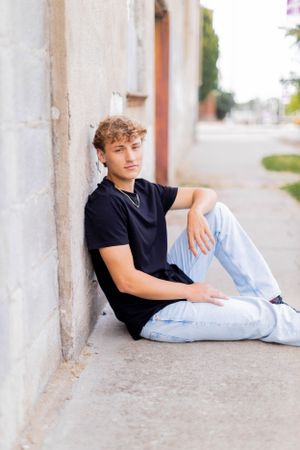 Senior session with boy in casual wear sits on ground against white wall in Springfield, Missouri.
