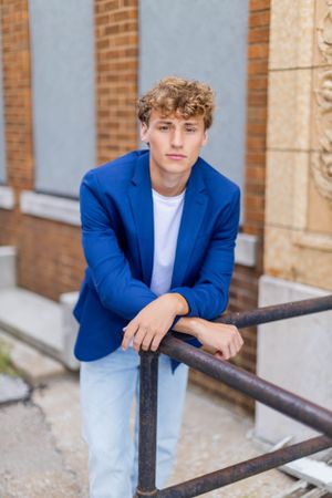 Senior session with boy in blue blazer and light jeans leans on railing by brick building in Springfield, Missouri.