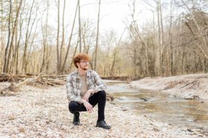 Senior session with boy in plaid shirt crouches by stream in winter forest landscape in Springfield, Missouri.