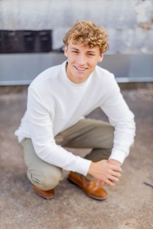 Senior session with boy in white sweater and khakis crouches down while wearing brown shoes outdoors in Springfield, Missouri.