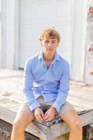 Senior session with boyin blue striped shirt and khaki shorts sits on wooden platform in casual summer pose in Springfield, Missouri.
