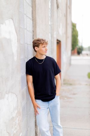 Senior session with boy in black t-shirt and light jeans poses against white wall outdoors in Springfield, Missouri.