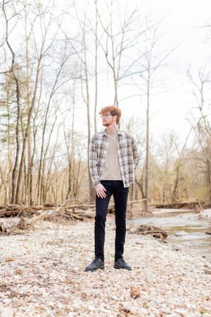 Senior session with boy in plaid shirt and dark jeans stands in wooded area near stream in Springfield, Missouri.