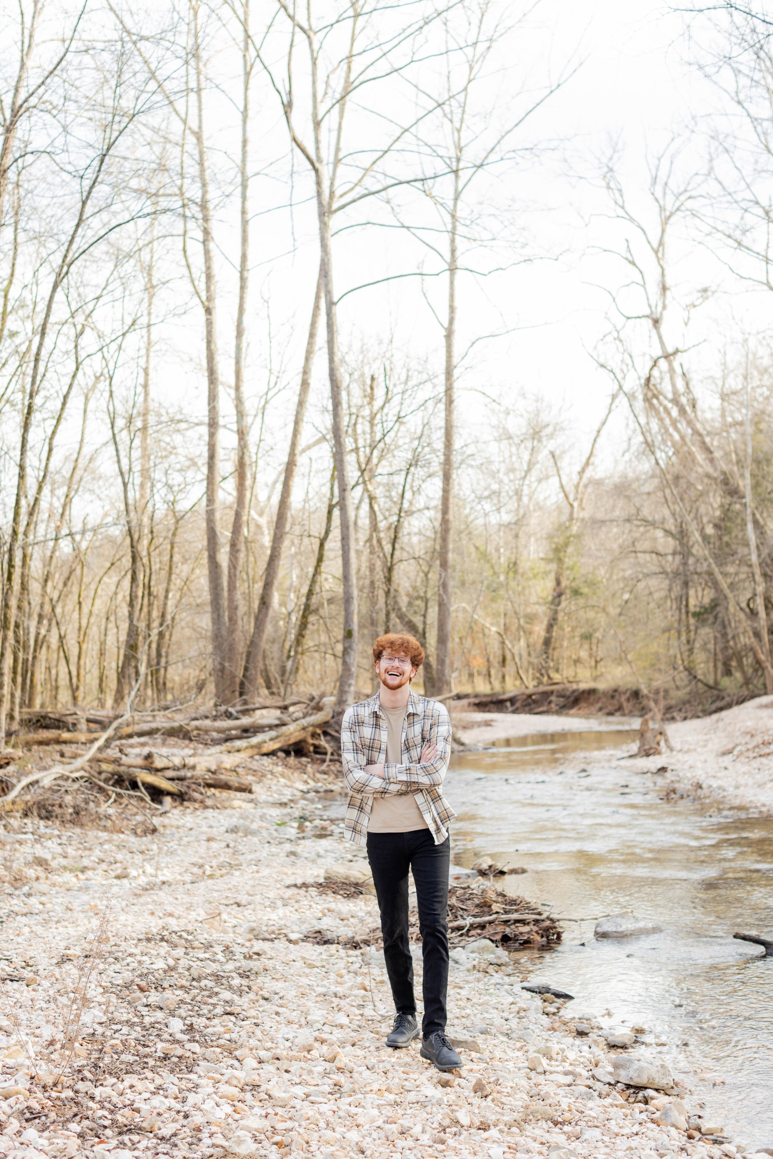 Senior session with boy wearing plaid shirt stands by flowing stream in winter forest landscape in Springfield, Missouri.