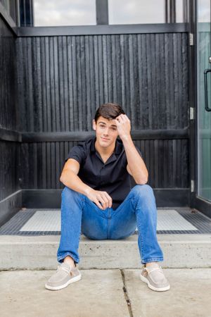 Senior session with boy in navy polo and jeans sits casually against industrial metal wall background in Springfield, Missouri.