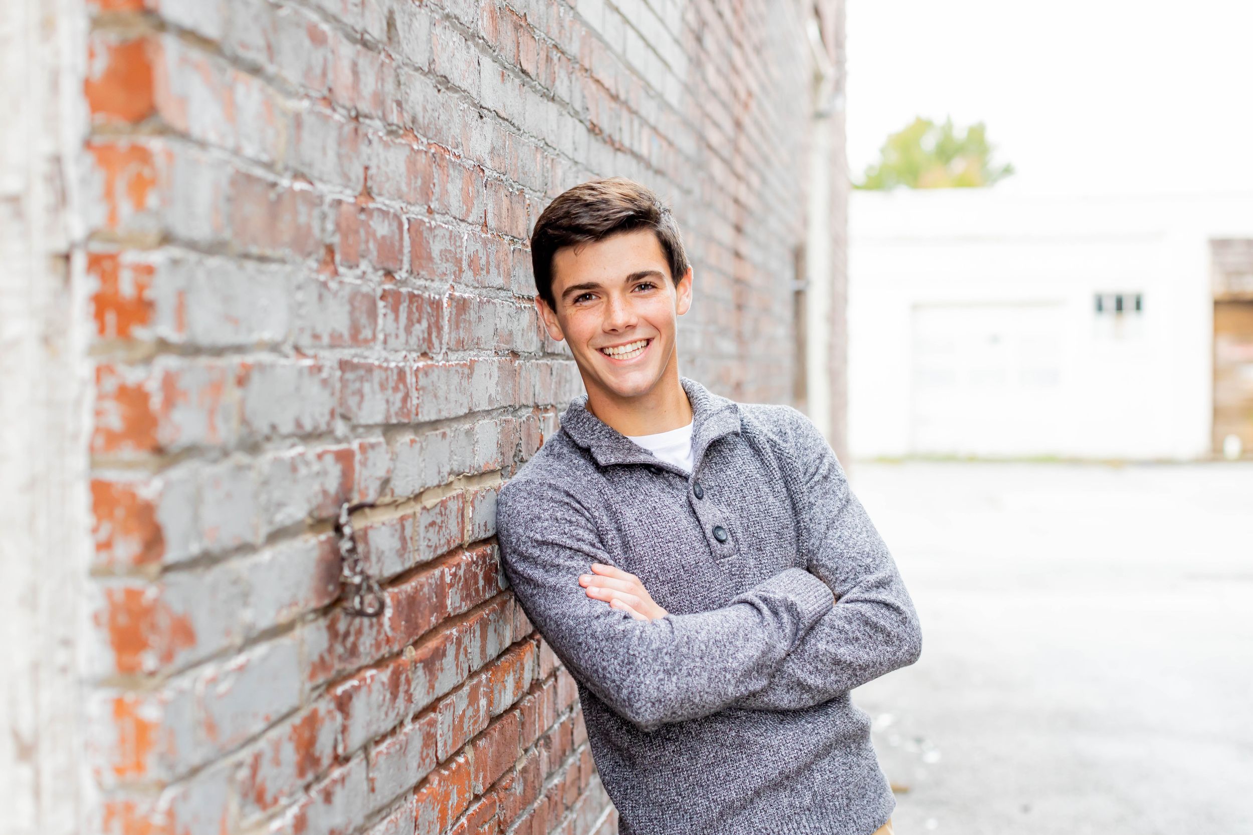 Senior session with boy in gray sweater leans against brick wall with arms crossed in Springfield, Missouri.