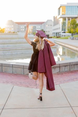 Graduate in black dress and burgundy coat twirling joyfully by a reflecting pool at sunset in Springfield, Missouri.