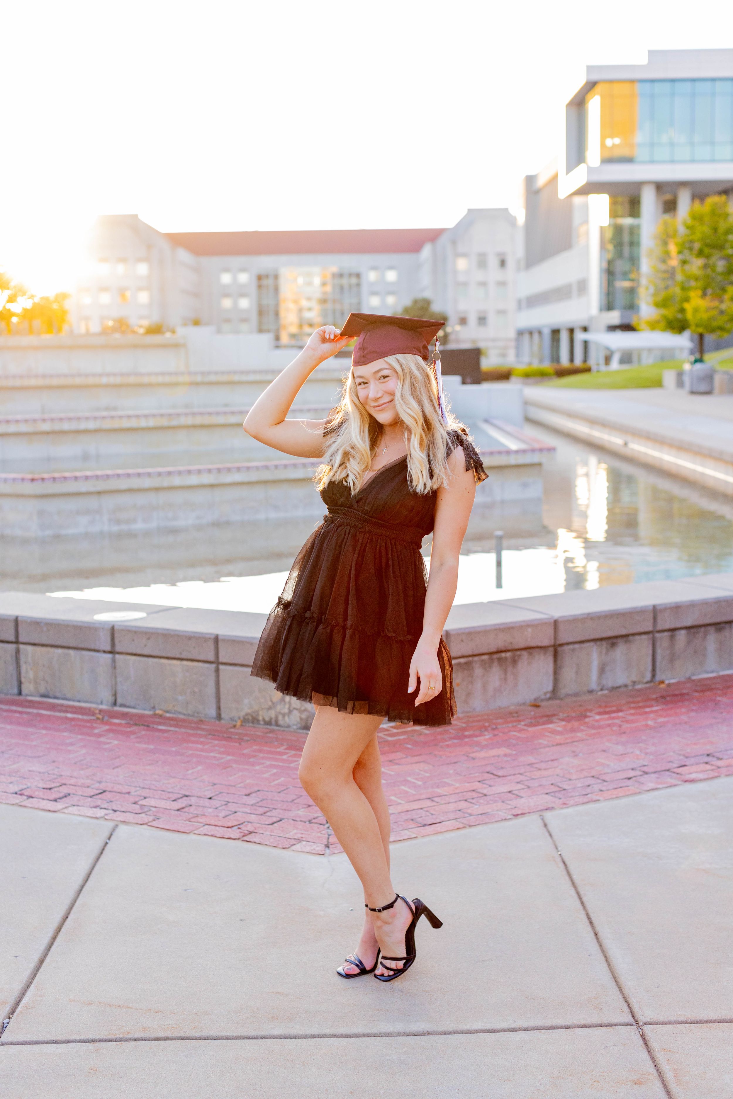 Graduate in black dress posing with cap near modern campus building during golden hour in Springfield, Missouri.