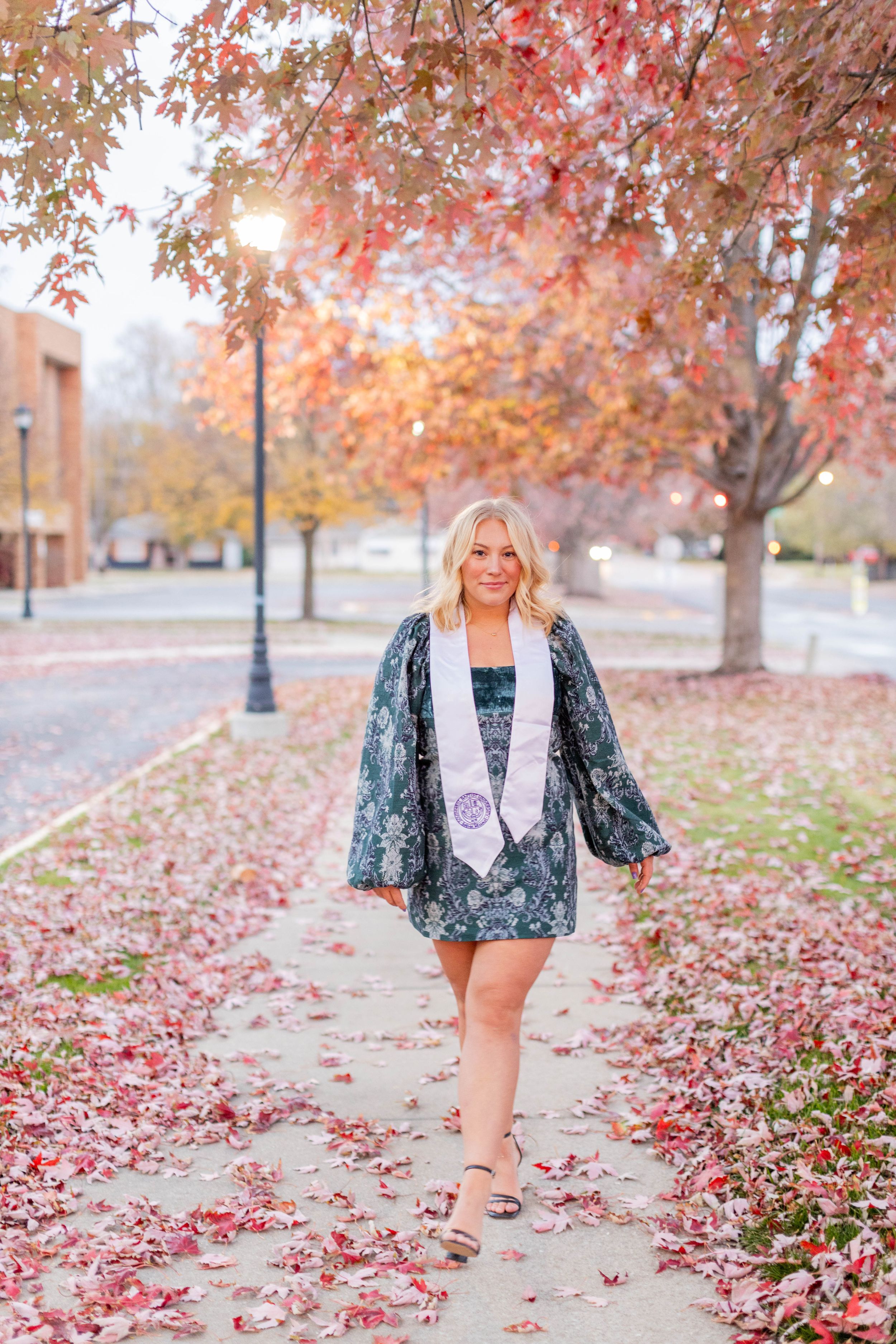 Graduate walking on path covered in pink fall leaves wearing patterned dress and graduation sash in Springfield, Missouri.
