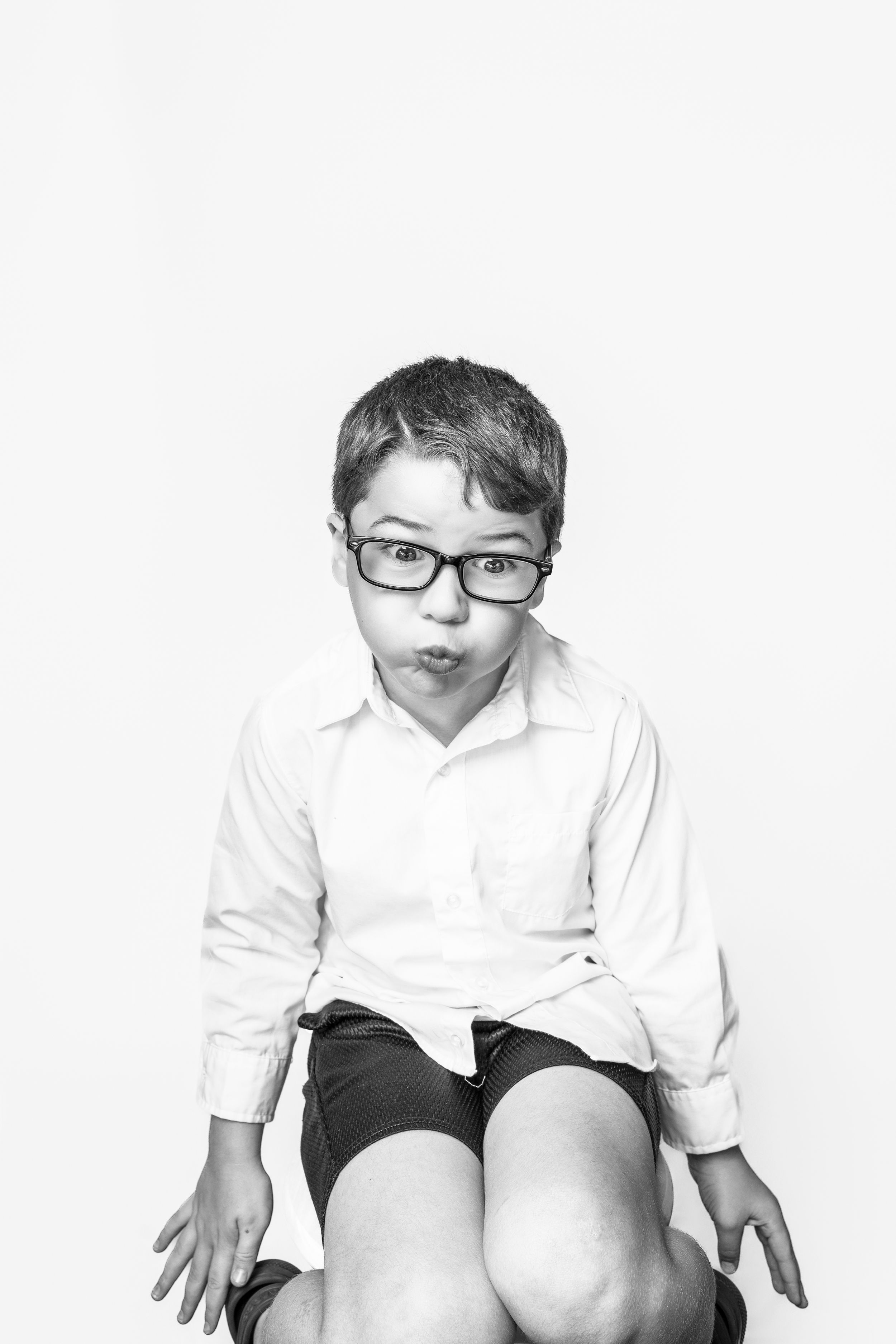 Black and white portrait of a child in glasses and white shirt making a playful expression against white background, Cocord, NC.
