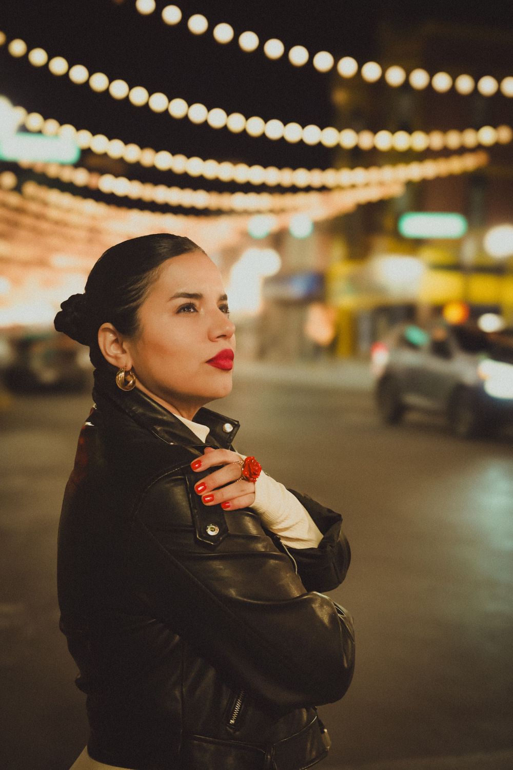 Woman in leather jacket stands confidently under string lights at night.