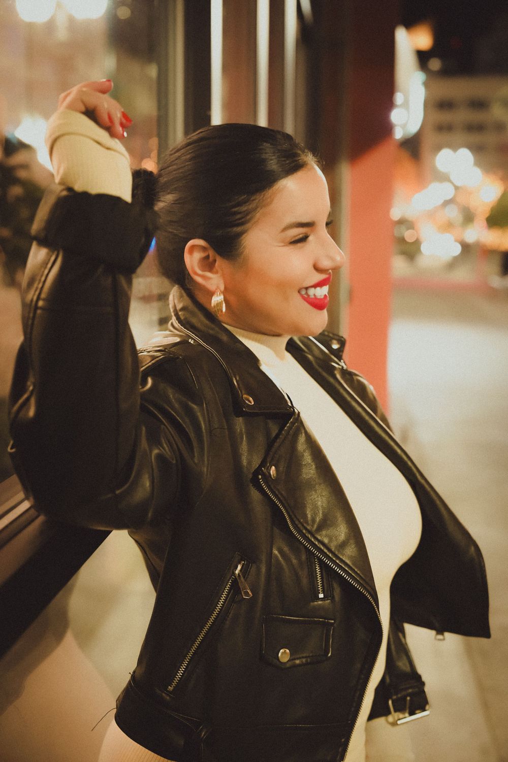 Woman in leather jacket and white top strikes confident pose against evening city backdrop.