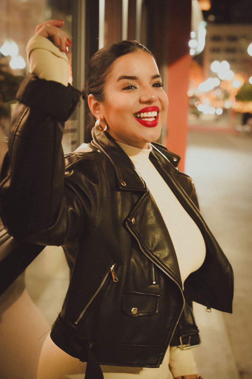 Woman in leather jacket and white top smiles joyfully while posing outdoors.