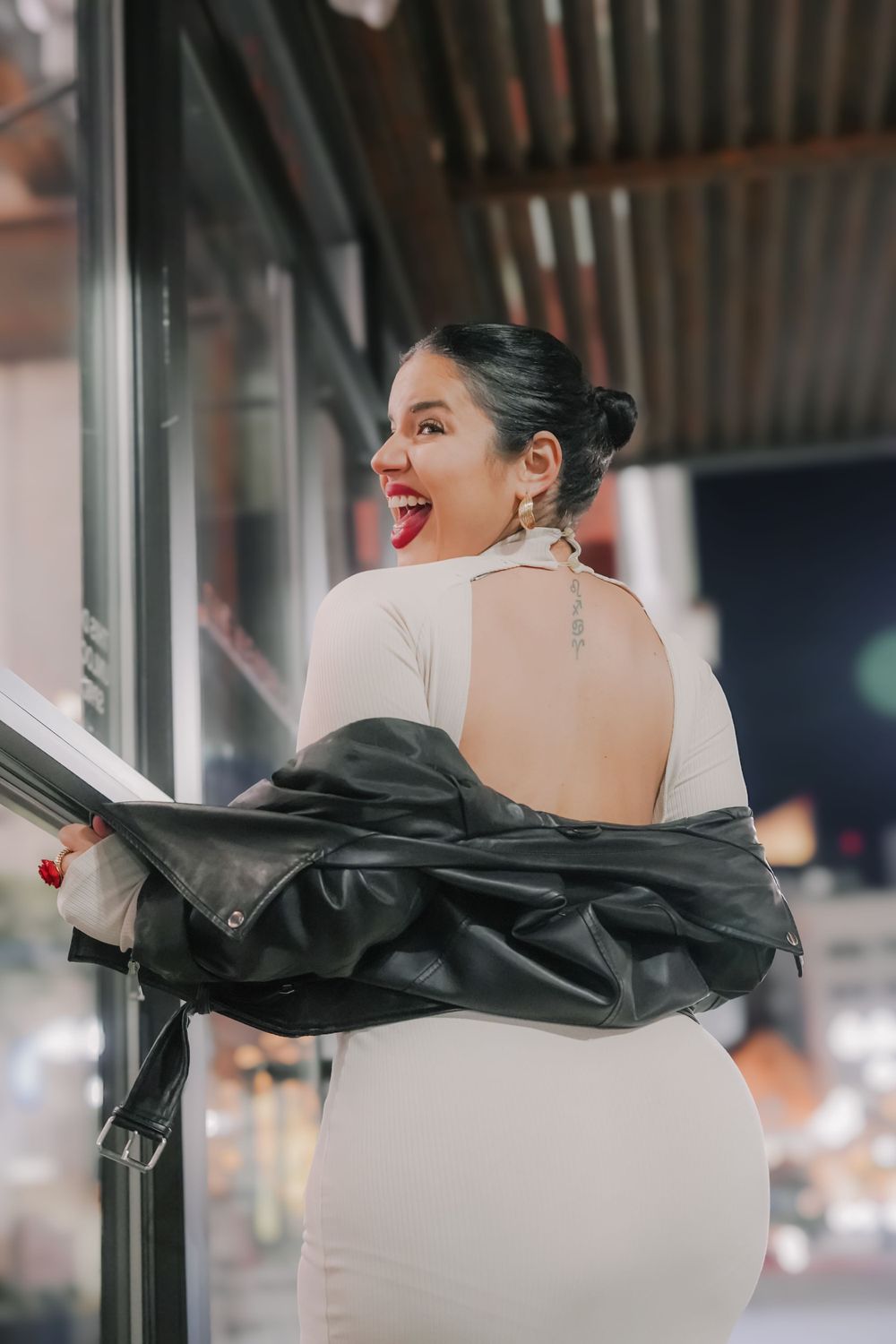 Person in white dress and leather jacket poses playfully showing bright red lipstick.