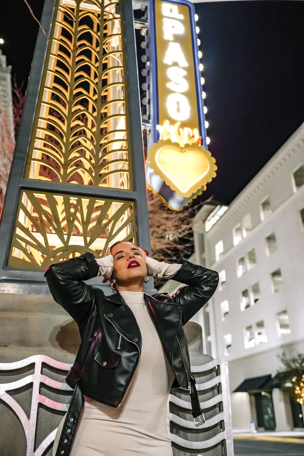 Person in leather jacket poses exuberantly beneath illuminated theater marquee at night.