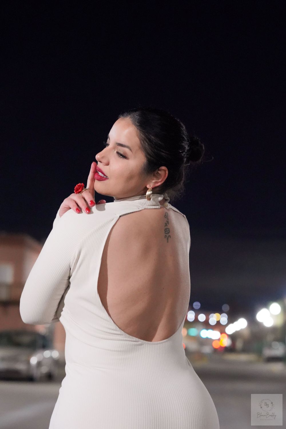 Person in white backless dress poses under night sky with city lights in background.
