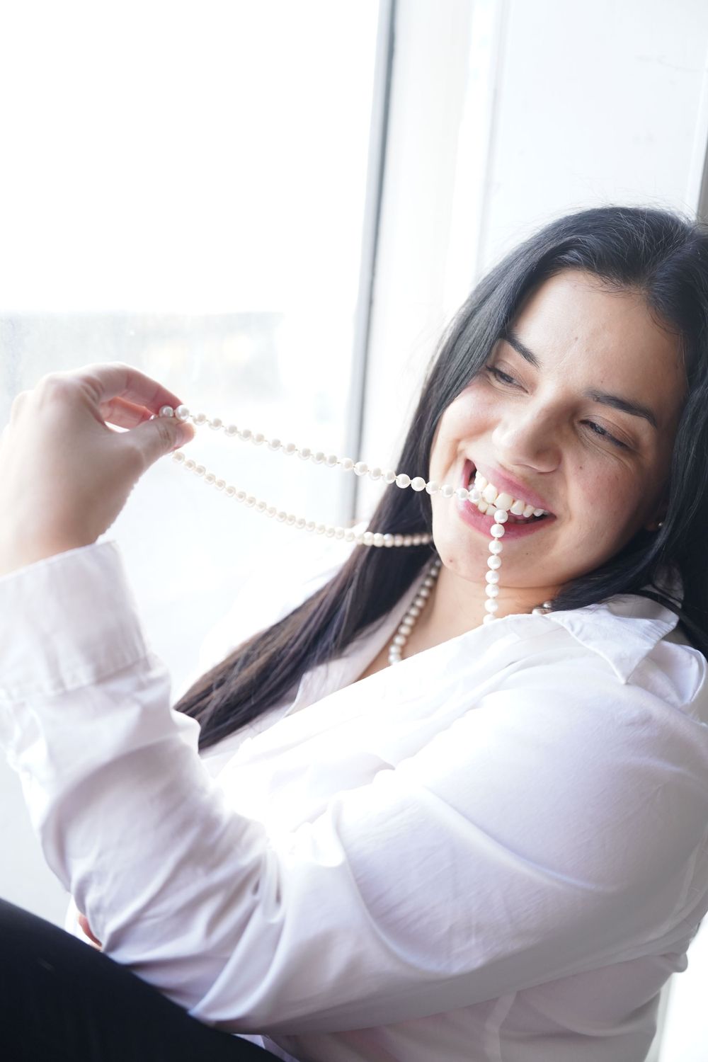 Person playfully poses with pearl necklace by bright window.