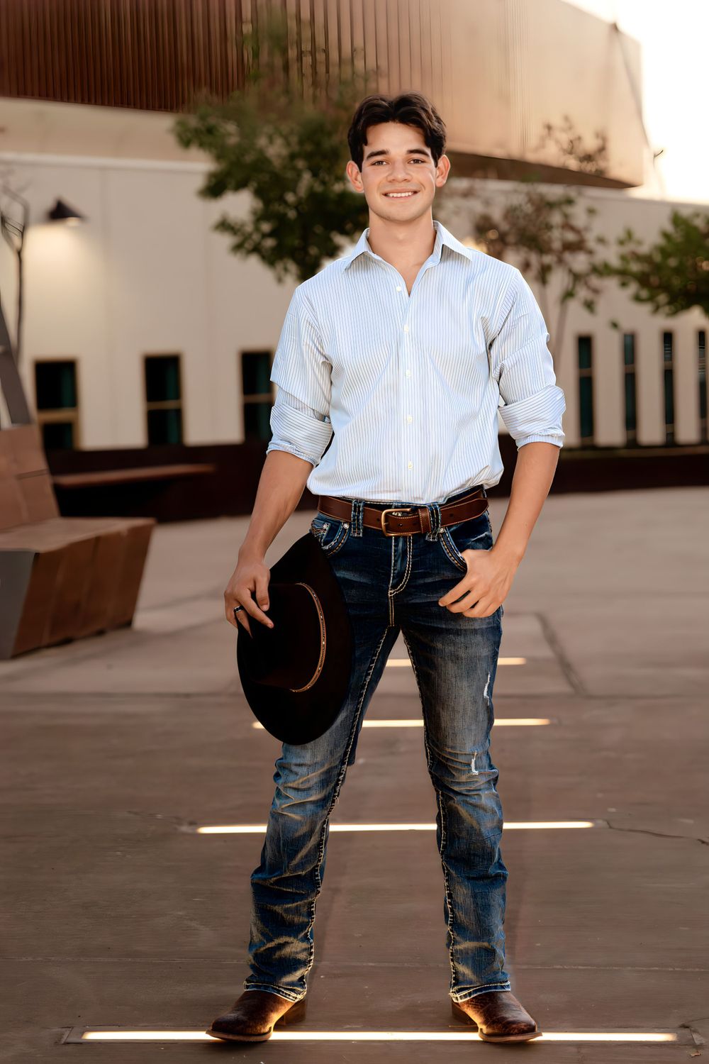 Person in western attire poses with cowboy hat against urban backdrop.
