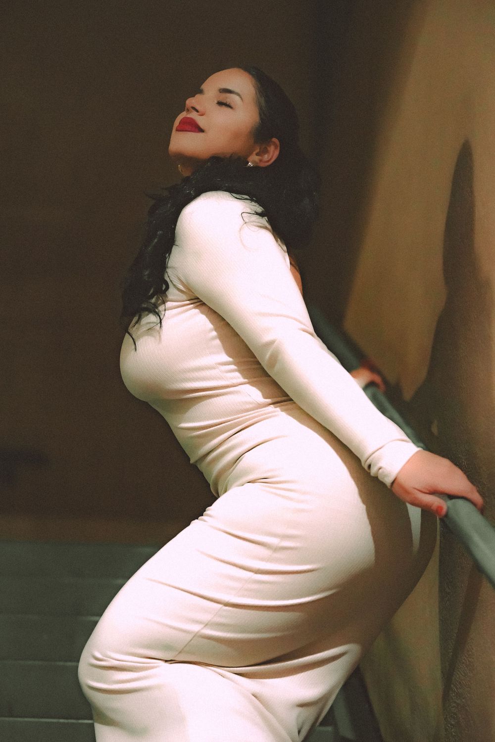 Woman in form-fitting white dress poses elegantly against wall in dramatic lighting.