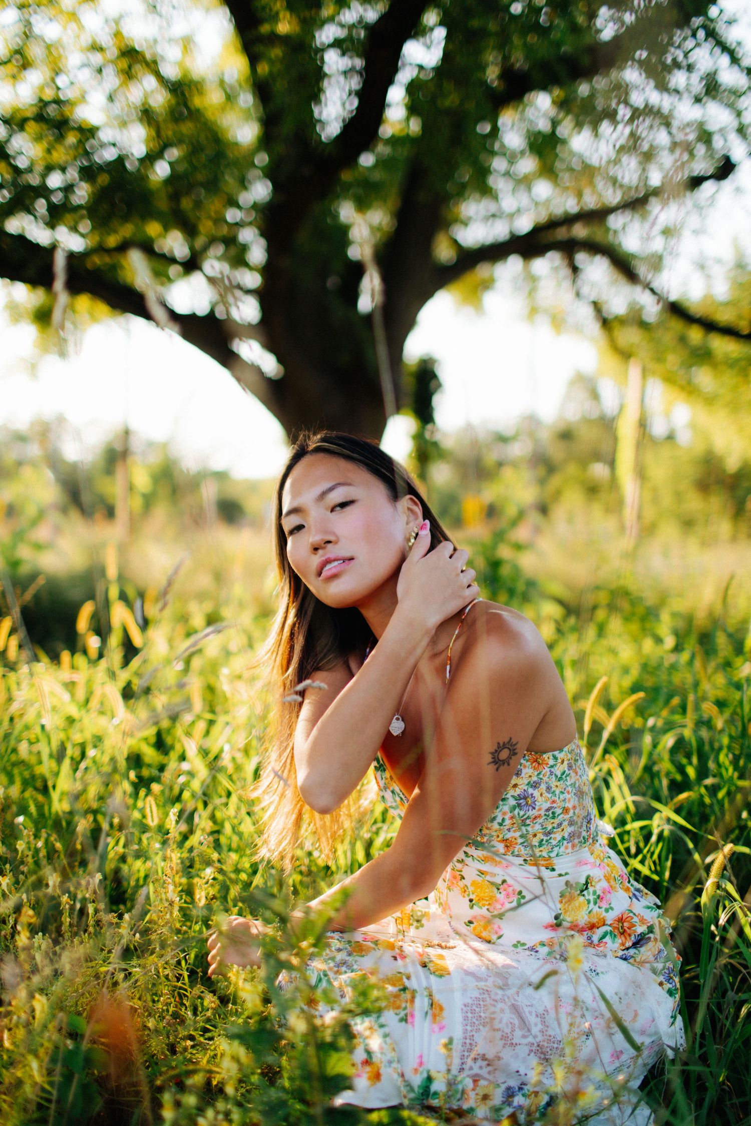 Person in floral dress poses elegantly in sunlit field with bokeh effect.