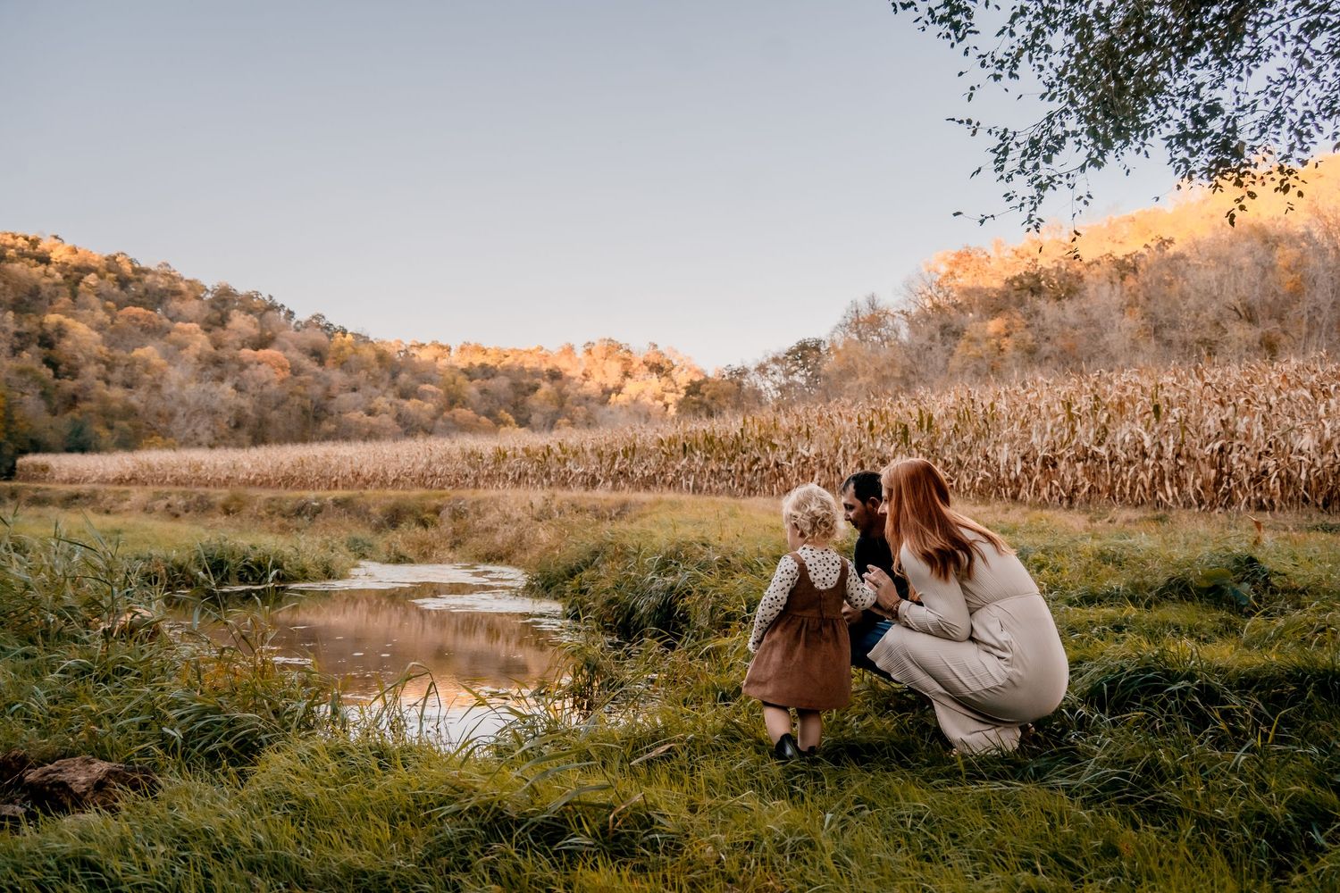 Adult and child crouching by stream at sunset in autumn landscape.