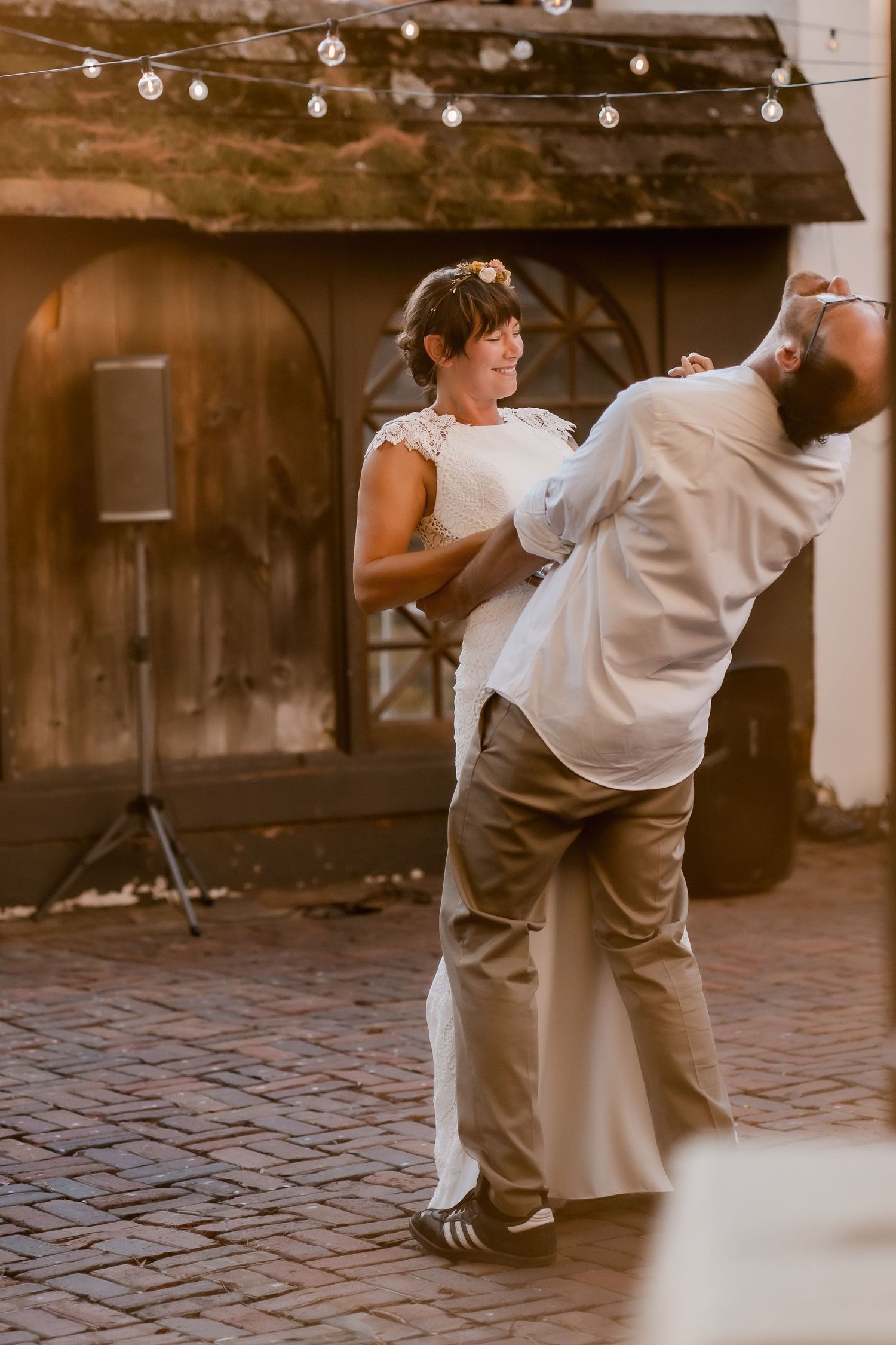 Wedding couple dances playfully on a rustic brick patio under string lights.