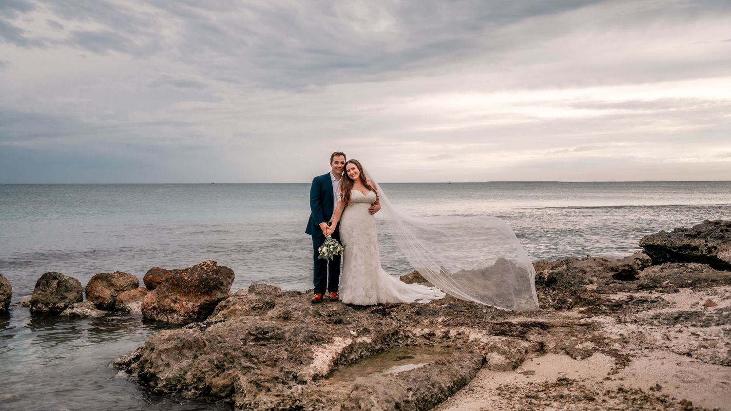 Newlywed couple posing on rocky beach with dramatic cloudy sky backdrop.