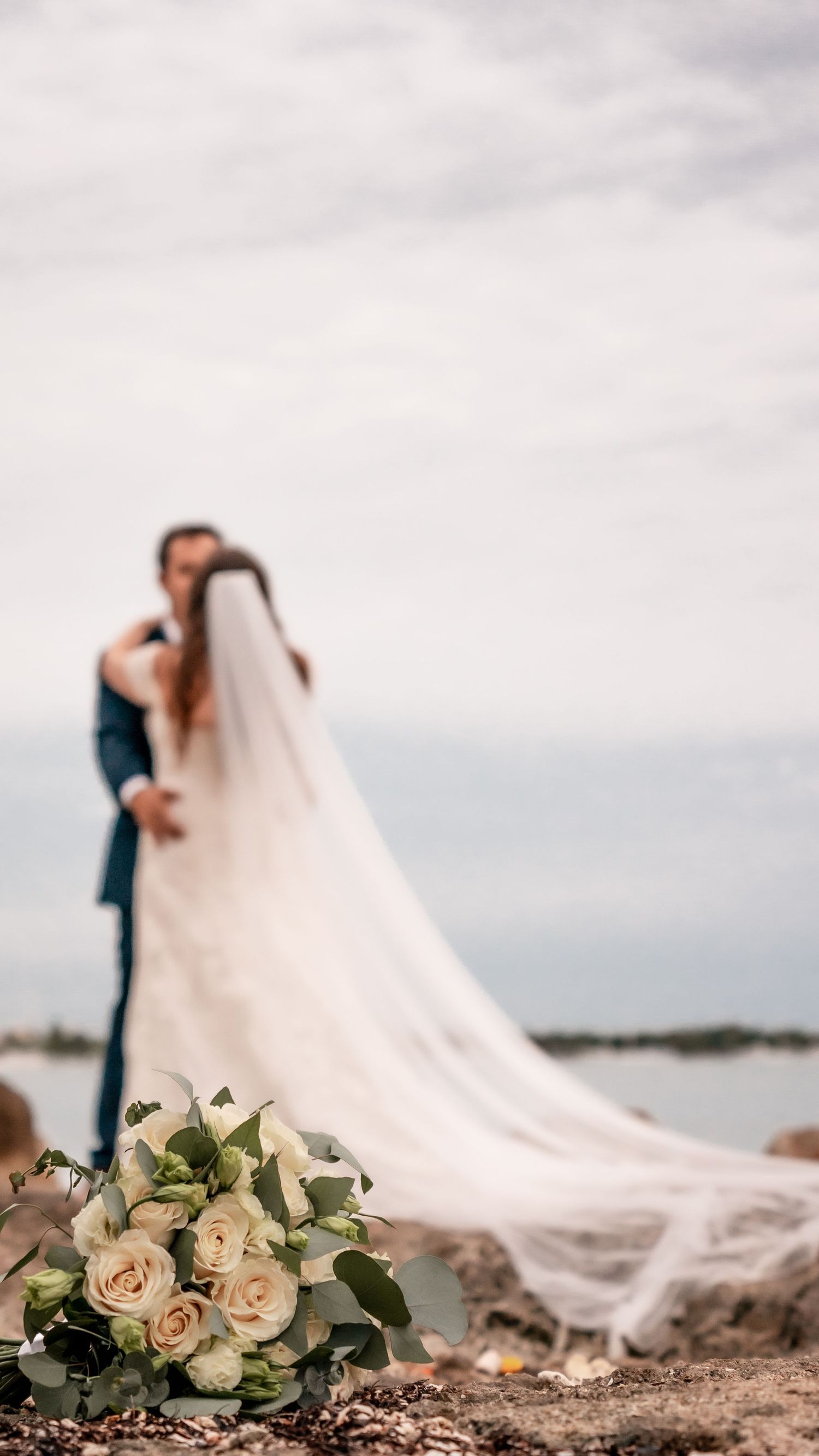 Wedding bouquet of roses in foreground with couple silhouetted against ocean view.