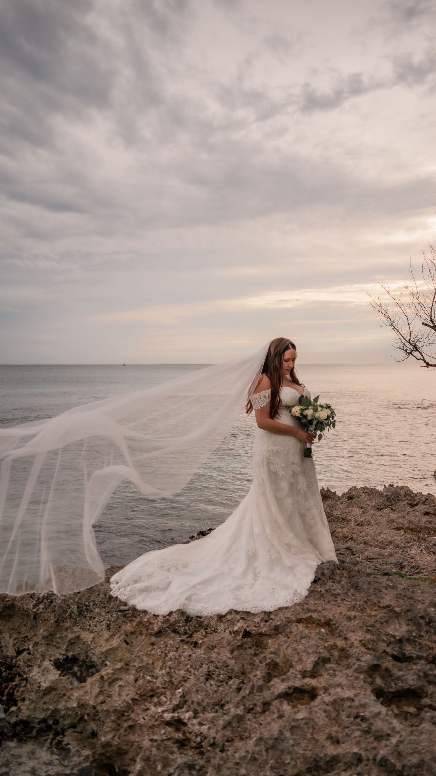 Bride in flowing white dress poses dramatically on rocky oceanside cliff.