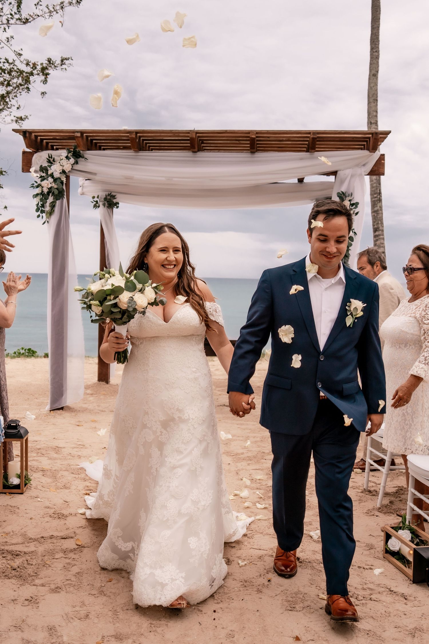 Newlyweds walk joyfully under white canopy during beach wedding ceremony.
