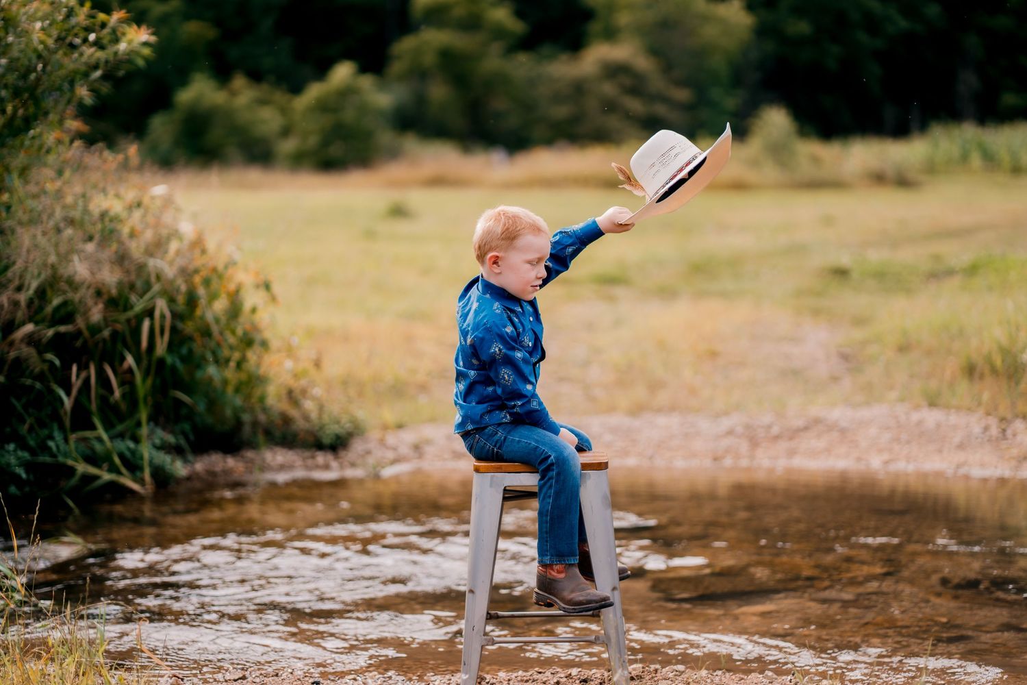 Child in blue sitting on stool by stream holding white hat in countryside setting.