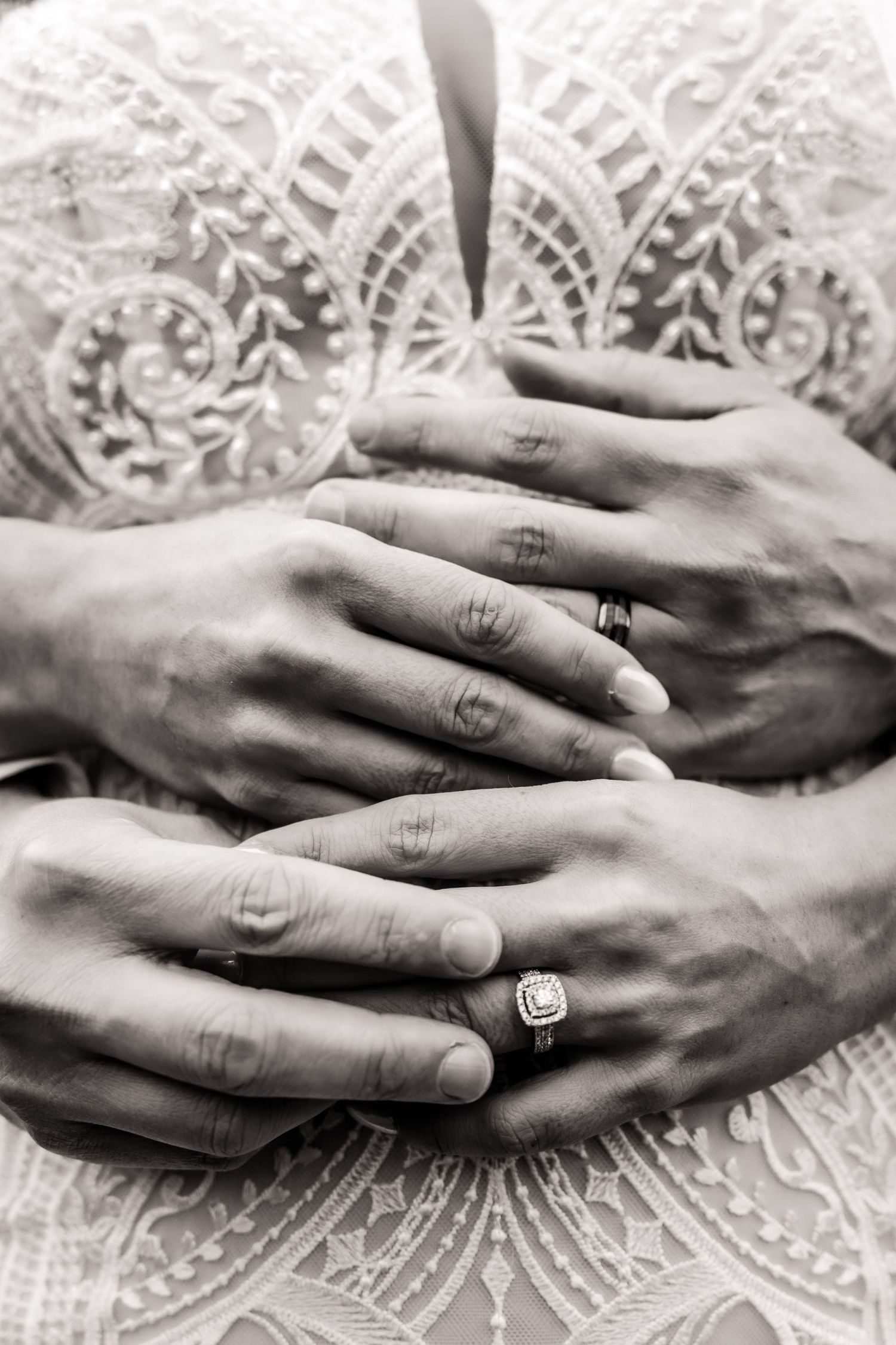 Black and white close-up of hands with wedding rings intertwined on lace fabric.