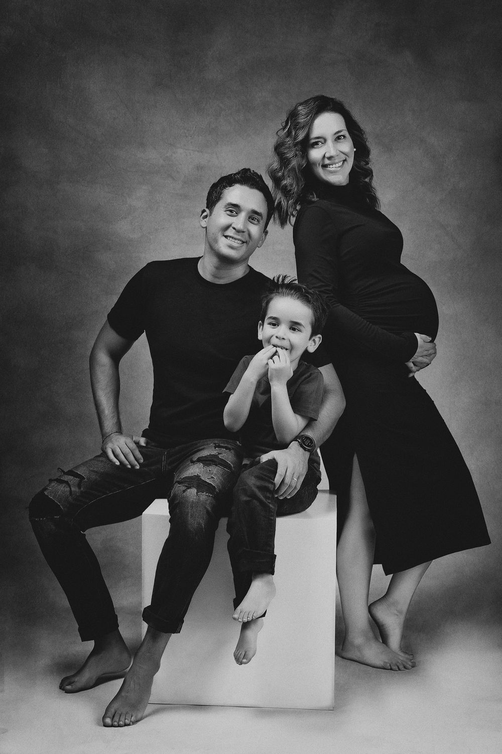 Black and white maternity photo of smiling family with young boy in studio setting.