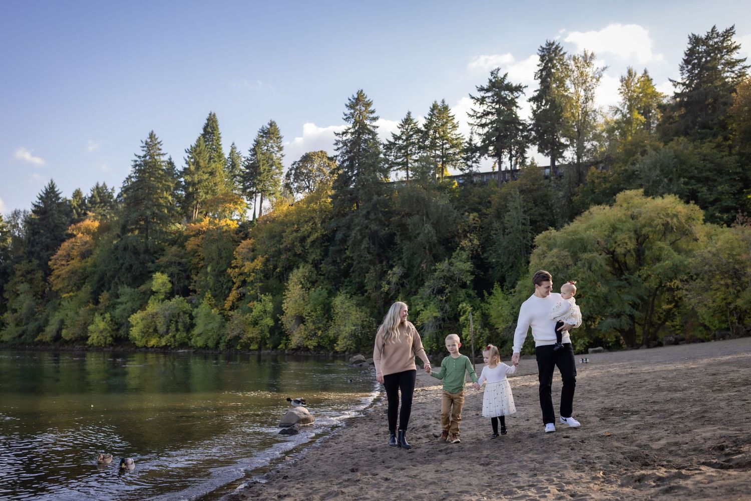 Family of five walking hand in hand along a scenic riverbank surrounded by tall trees.