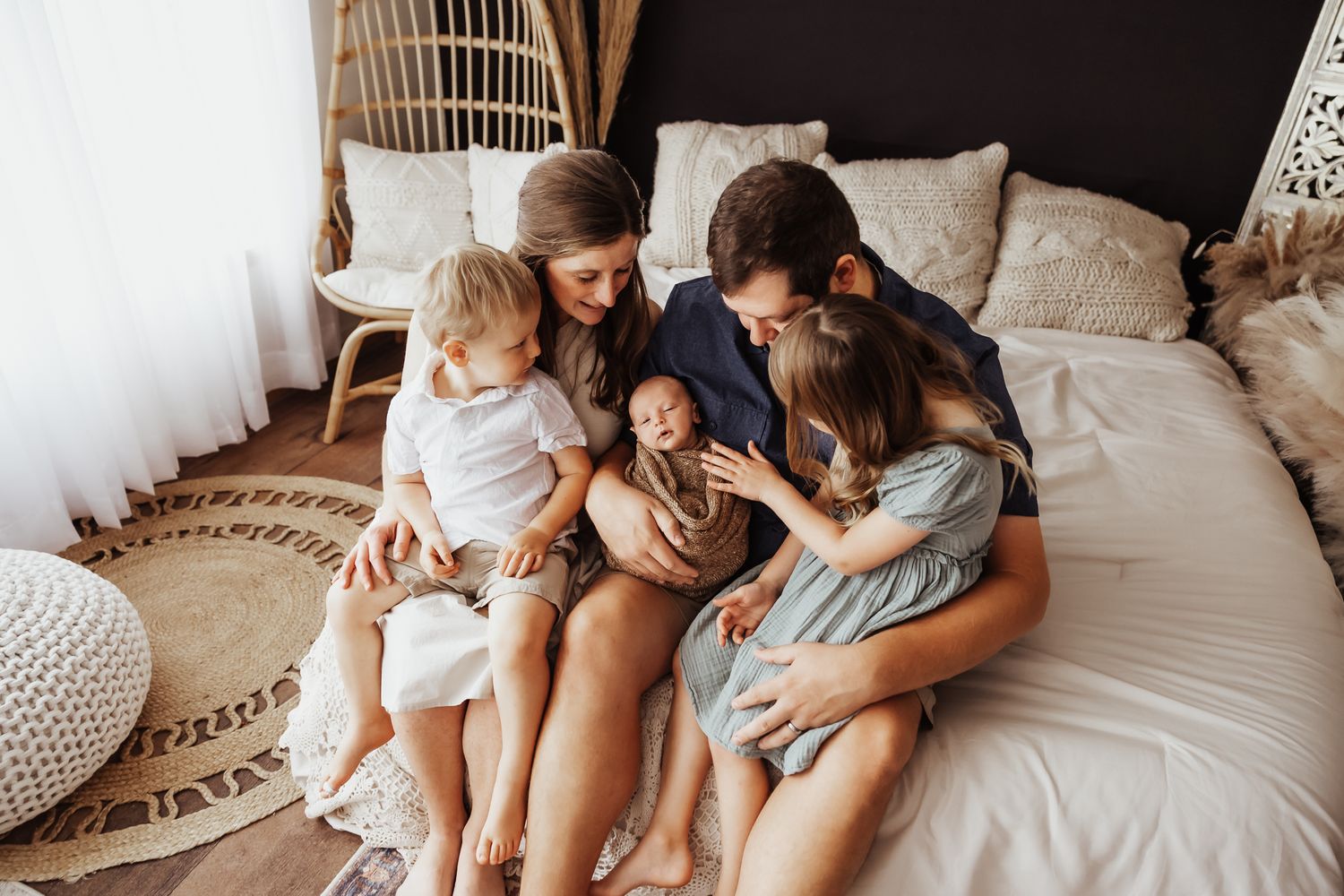 Happy family of five sitting on bed together admiring their newborn baby wrapped in tan swaddle.