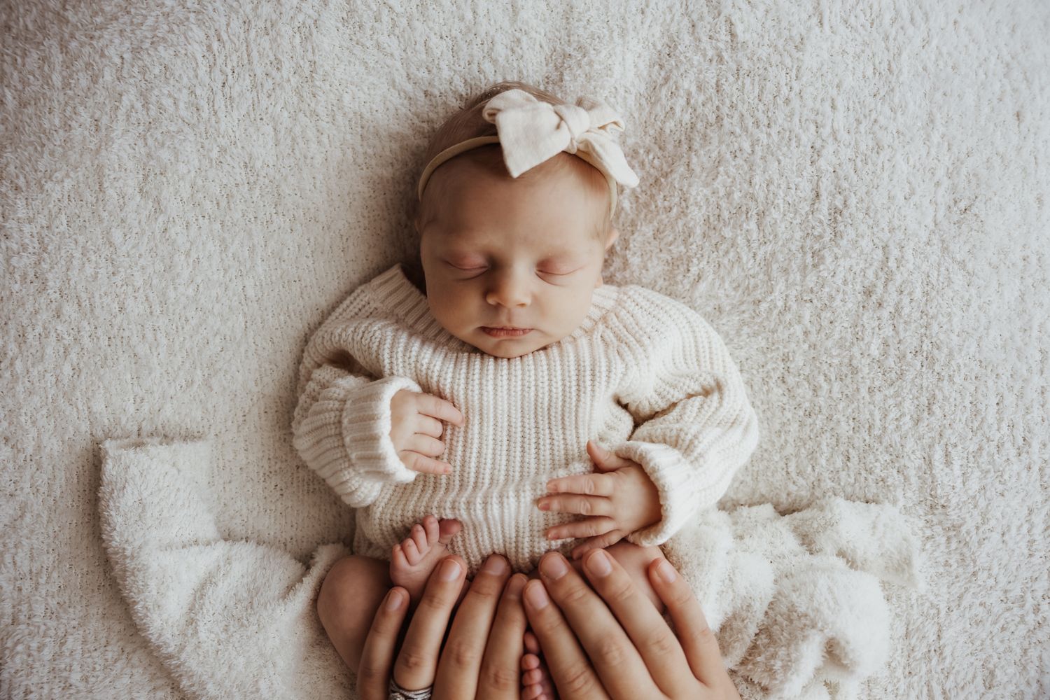 Sleeping newborn baby in cream knit sweater with bow headband held by multiple hands on white fur.