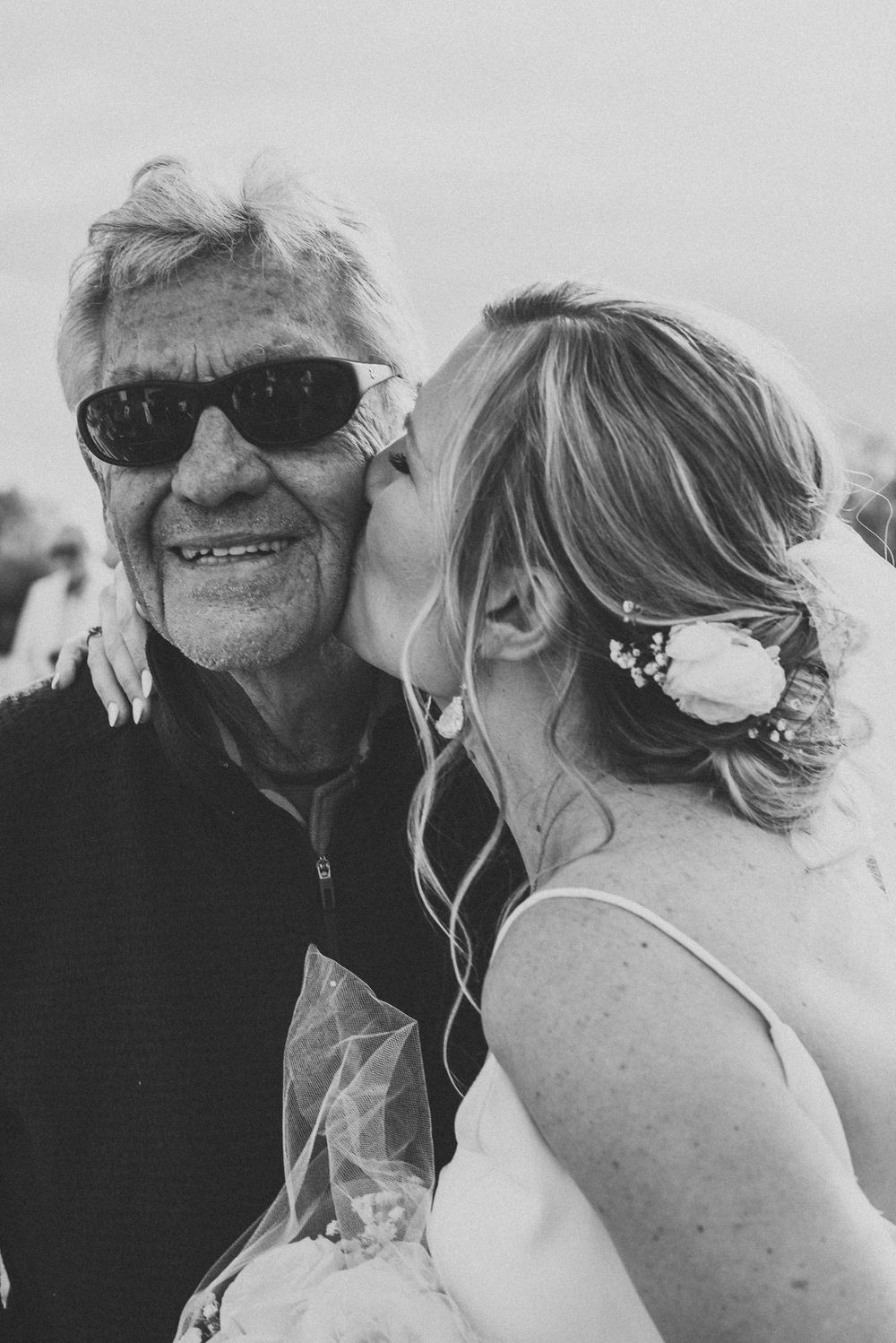 Black and white photo of a bride kissing an elderly man wearing sunglasses at a wedding celebration.
